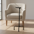 Black quad cane with soft gray ergonomic grip standing upright on pale oak floor beside a neutral armchair