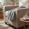 Soft fog-gray heated lap blanket folded on the arm of a beige fabric recliner with a cup of tea on a wooden side table